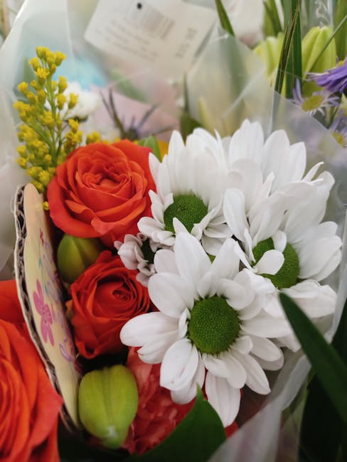 A vibrant floral arrangement featuring orange roses with tightly curled petals, white daisies with bright green centers, and sprigs of yellow solidago, all wrapped in clear cellophane and floral paper
