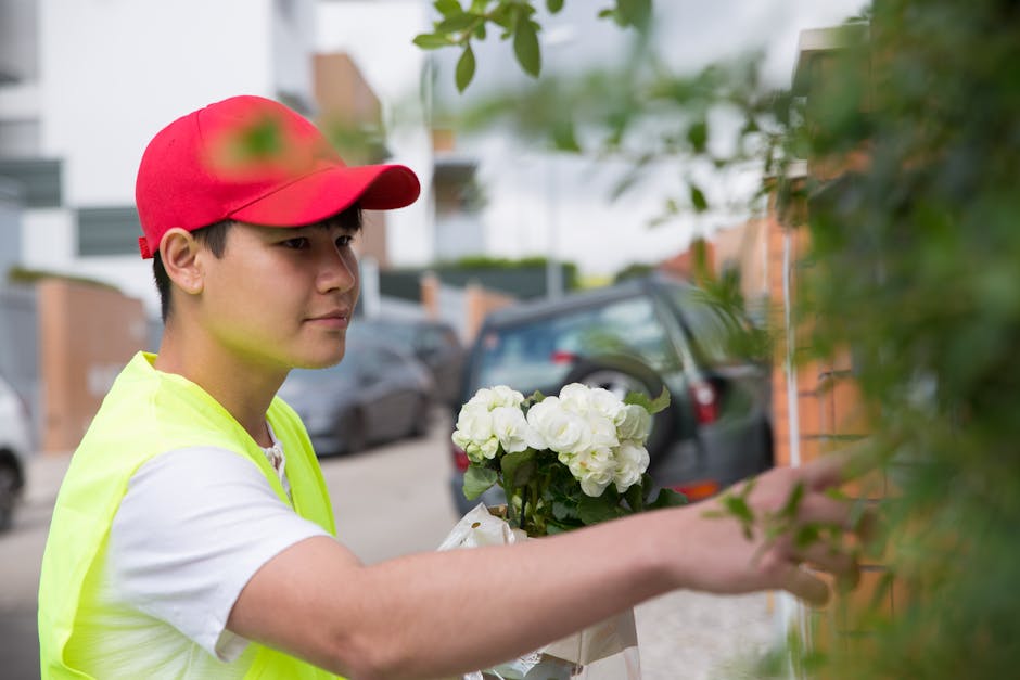A delivery person, wearing a bright yellow high-visibility vest and a red cap, is seen placing a floral gift consisting of white hydrangeas into a green leafy bush. The individual has a focused expres