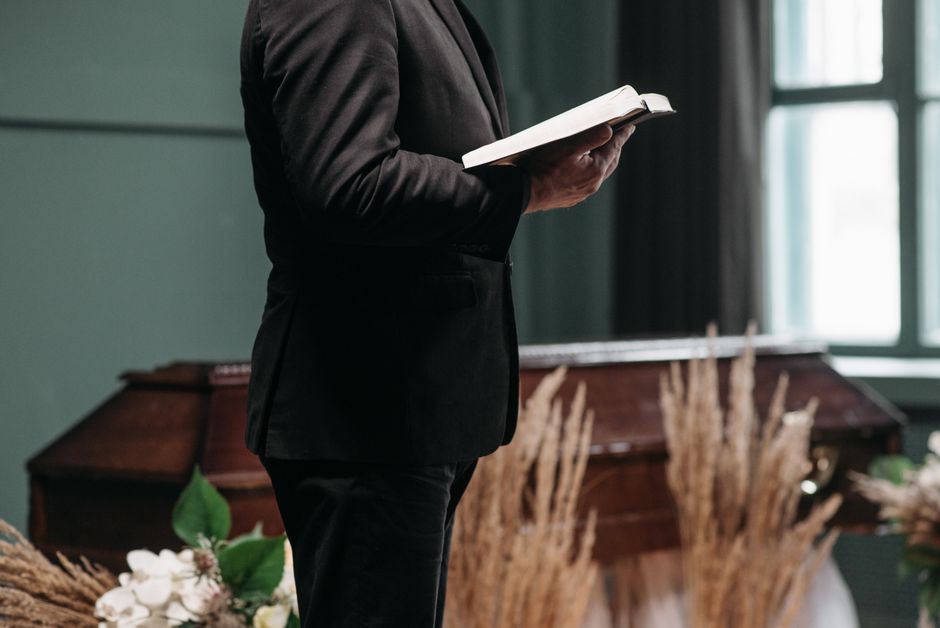 A person dressed in a black suit holding an open book, standing indoors near a wooden funeral casket decorated with dried pampas grass and white flowers. The scene is softly lit with natural light com