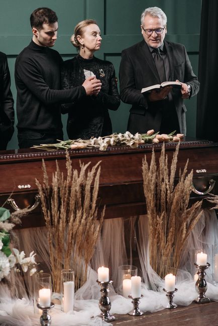 A funeral floral arrangement at a memorial service, featuring cream and white flowers including roses, lilies, and carnations, set on a polished wooden casket adorned with beige and white linen. Tall 