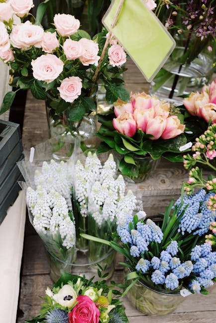 A display of fresh flower arrangements, including light pink roses in a tall glass vase, pink tulips with green leaves in a round bouquet, white grape hyacinths wrapped in clear plastic, and clusters 
