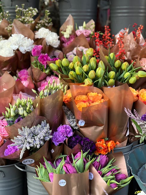 A vibrant display of fresh flowers arranged in brown craft paper and clear wrapping, including white and purple carnations, pink and purple tulips, orange roses, and green tulip buds. The bouquets are
